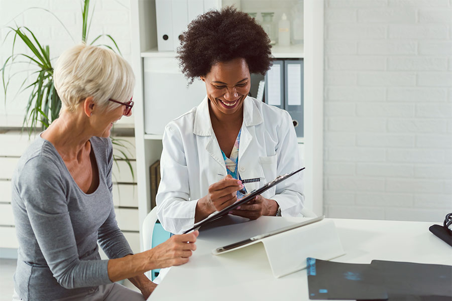 Employee Benefits - Portrait of a Smiling Doctor Sitting in a Medical Office with a Senior Woman While Holding a Tablet in Her Hands