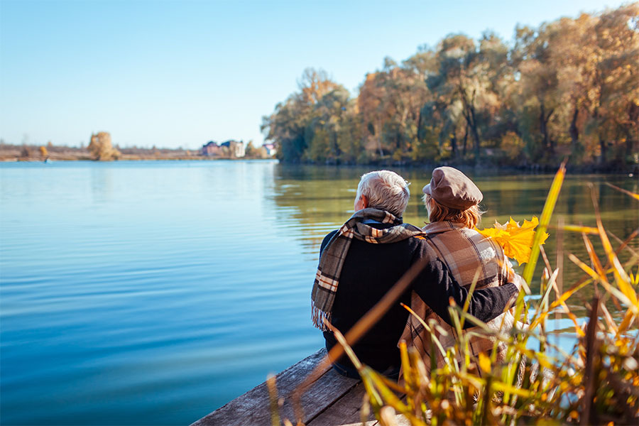 Health Insurance - Rear View of an Elderly Couple Sitting and Cuddling Each Other By a Calm Lake During the Fall